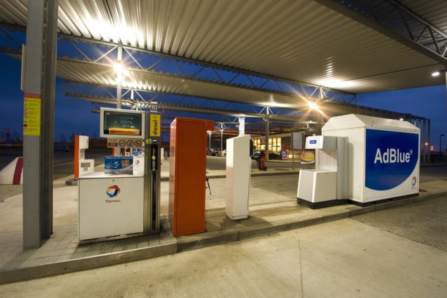 Vue d'un îlot carburant réservé aux camions, placé sous le auvent de la station autoroutière atypique de Hellebeq, en Belgique. A côté du volucompteur, une borne pour le gonflage des pneumatiques et plus à gauche un réservoir Ad Blue. A l'arrière plan, la boutique Bonjour. Prise de vue réalisée de nuit, avec les différents éclairages de la station actifs.